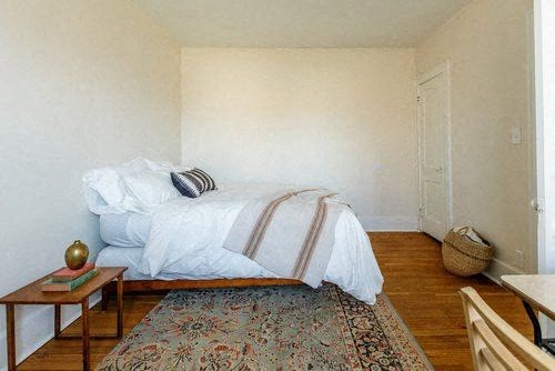 A bedroom with a bed, a small table, a rug and a basket on the floor.