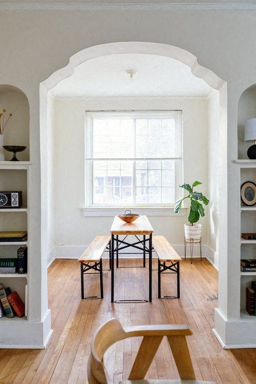 A table with chairs and a potted plant sits in a room with white walls and a window.