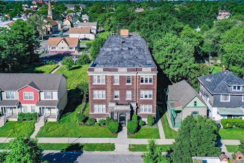 A large red brick building with a black roof is surrounded by green trees and smaller houses.