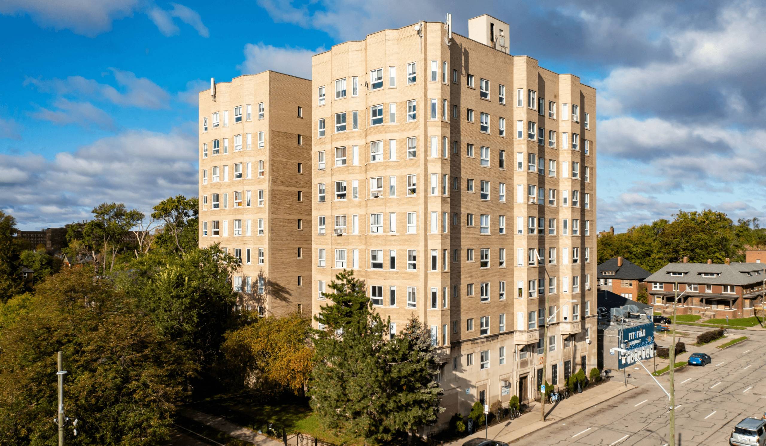 A large beige building with many windows is surrounded by trees and a street in front.