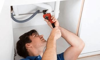 A plumber in a blue shirt is fixing a white pipe with a red tool.
