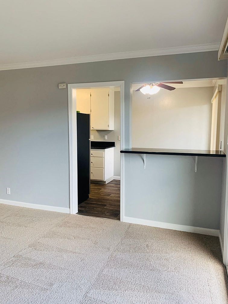 A room with a ceiling fan and a kitchen area in the background.