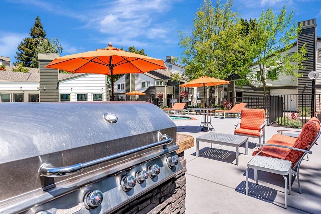 A silver grill is in the foreground of a patio with orange chairs and umbrellas.