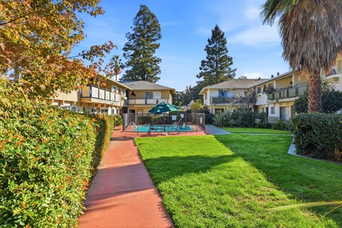 A pathway leads through a grassy area with a tennis court and apartment buildings in the background.