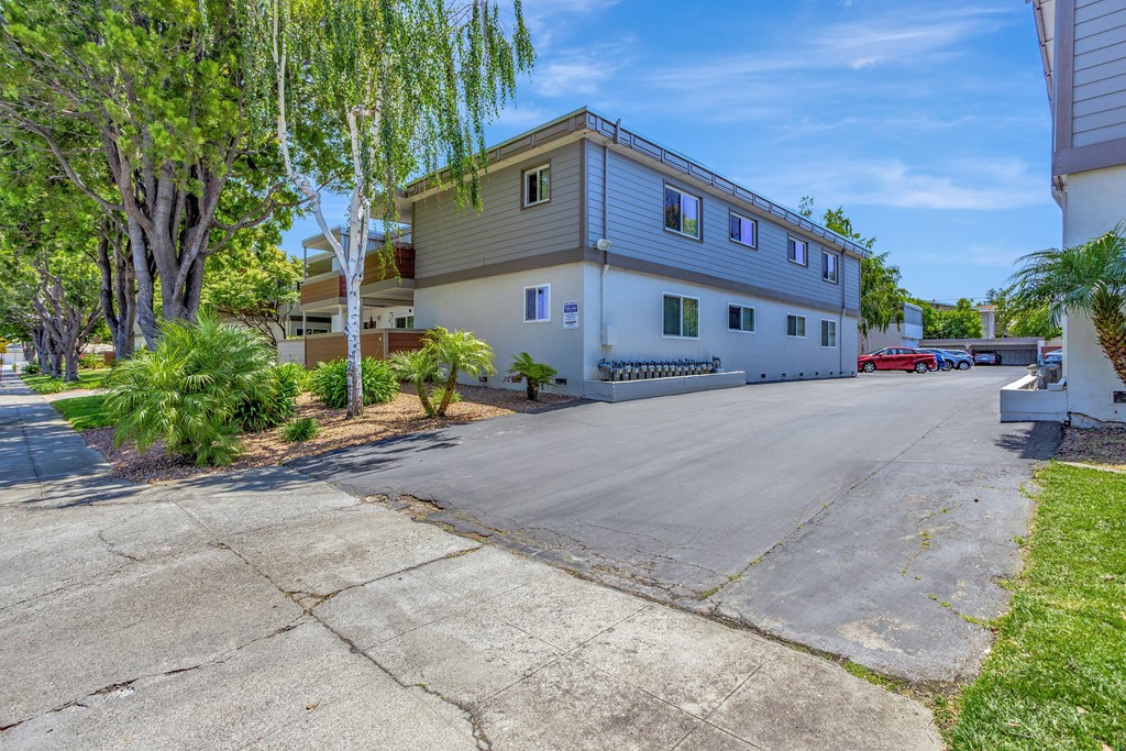 A two-story building with a white exterior and a grey roof is surrounded by trees and a driveway.