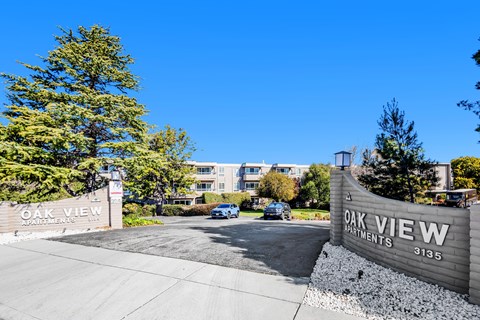 The image shows the entrance to the Oak View Apartments with a clear blue sky in the background.