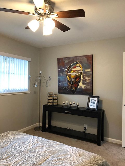 A bedroom with a bed, a ceiling fan, a painting, and a shelf with various items.