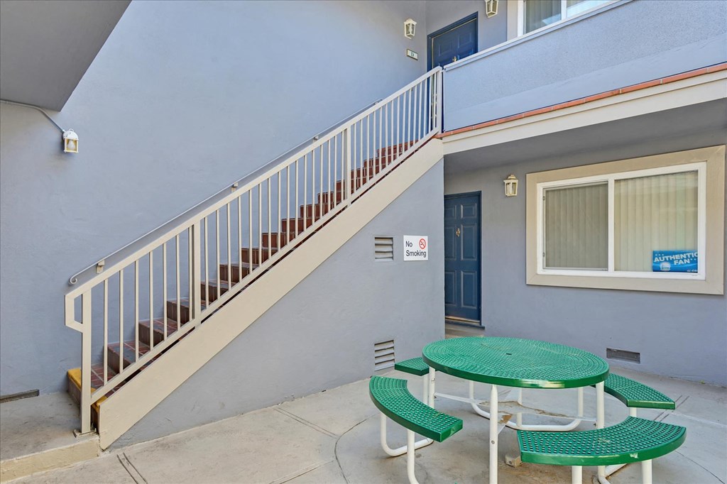A green table and chairs are on a patio outside a building with a staircase.