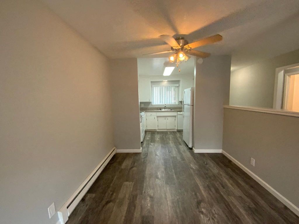 A room with a ceiling fan and a kitchen in the background.