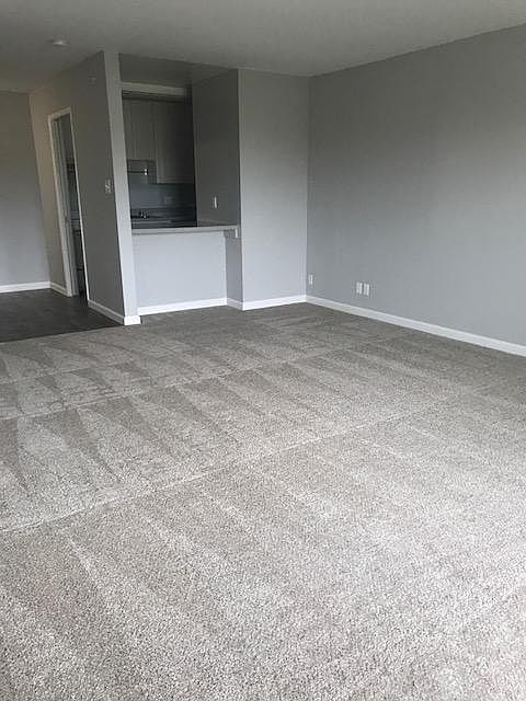 A room with a carpeted floor and a kitchen area in the background.