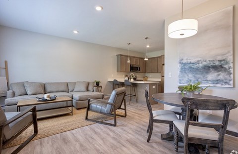 A modern living room with a grey sofa, a wooden dining table and chairs, and a kitchen area in the background.