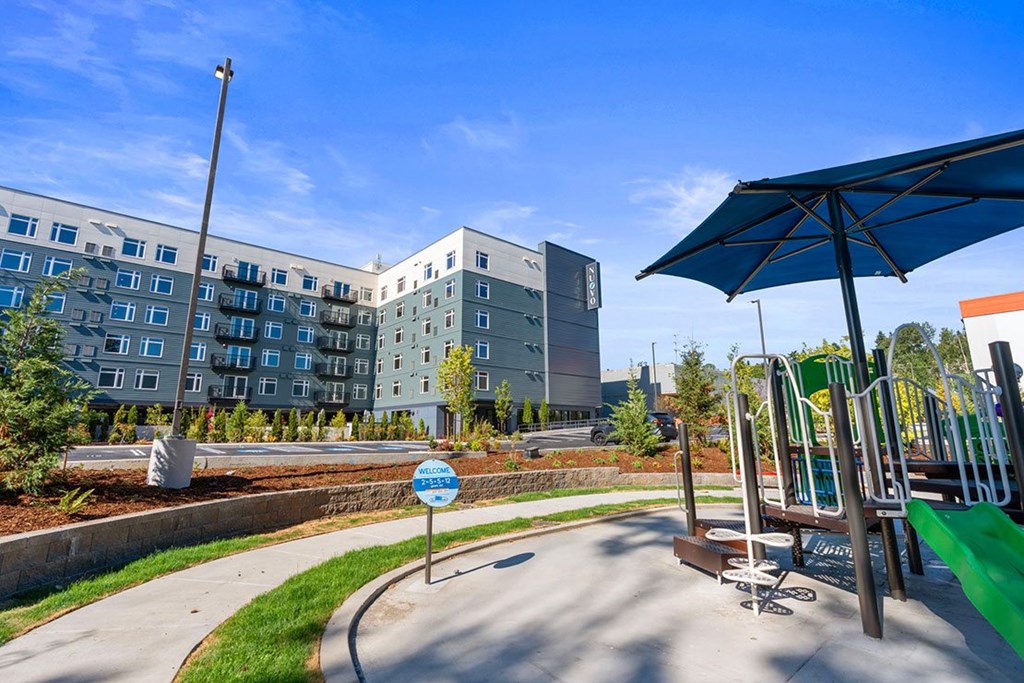 A playground with a blue sign in the middle of a paved area with a building in the background.