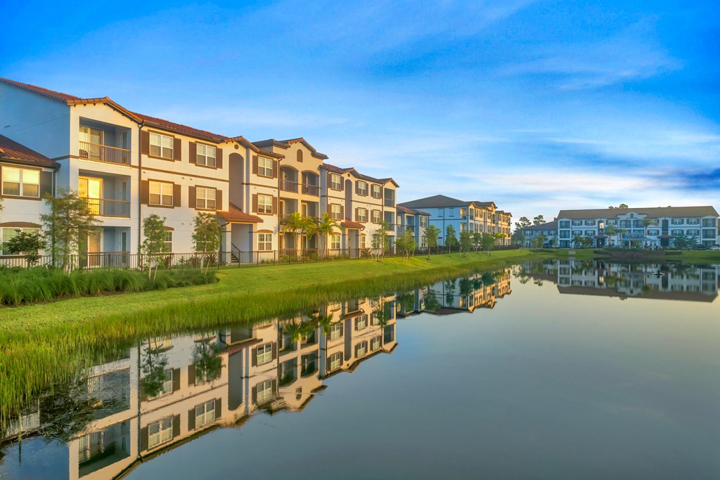 A row of modern apartment buildings are reflected in the calm water of a lake.