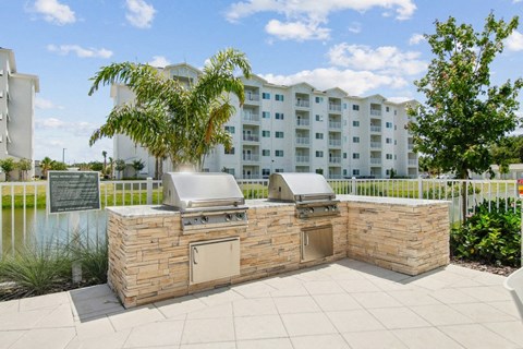 A stone barbecue is in the middle of a paved area with a building in the background.