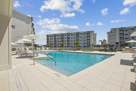 A swimming pool surrounded by lounge chairs and umbrellas with apartment buildings in the background.