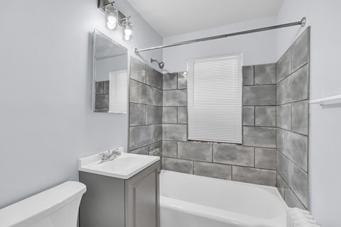 A bathroom with a white tub, sink, and grey tiled walls.