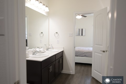 A bathroom with a double sink vanity and a bedroom with a bed and a ceiling fan.