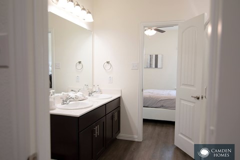 A bathroom with a double sink vanity and a bedroom with a bed and a ceiling fan.