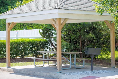 A wooden gazebo with a picnic table in front of a white fence and greenery.