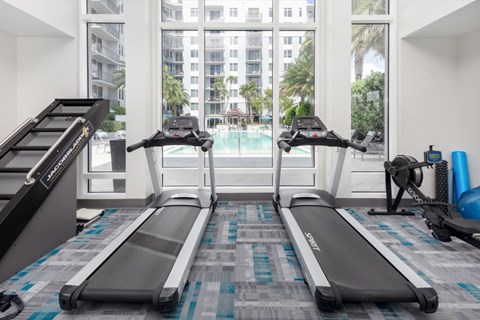 A treadmill and a stationary bike are in a gym with a view of a pool and buildings outside.