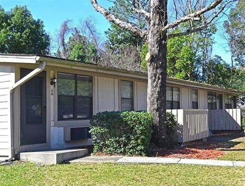 A house with a grey siding and a white door is surrounded by greenery.