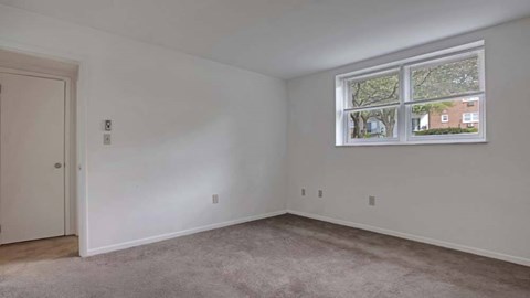 A room with a carpeted floor and a window showing a view of trees and houses outside.
