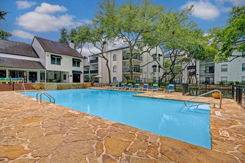 A swimming pool surrounded by stone and chairs in front of apartment buildings.