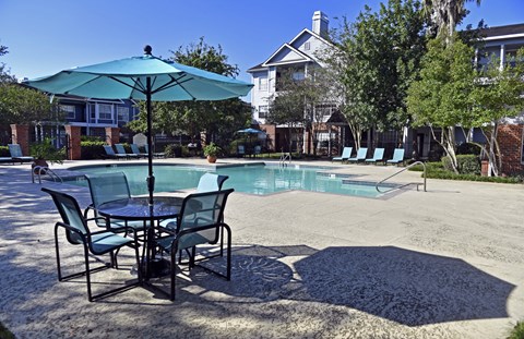 A table with chairs and an umbrella is in the foreground of a pool area.
