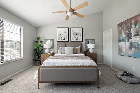 A bedroom with a bed, two lamps, a ceiling fan, and a painting on the wall.