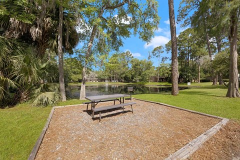 A picnic table sits in the middle of a gravel area with a grassy area and trees in the background.