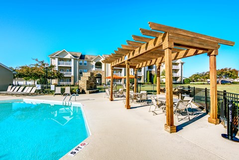 A pool with a pergola and chairs in front of apartment buildings.