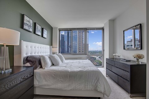 A bedroom with a large bed, a nightstand, and a window overlooking a cityscape.