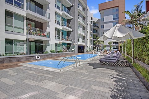 A swimming pool surrounded by a tiled patio and lounge chairs under a white umbrella.