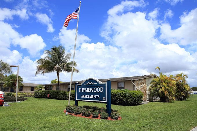 A sign for Thymewood Apartments is in front of a building with a flag on a pole.