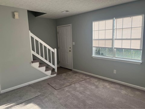 A room with a carpeted floor, a staircase, and a window with blinds.
