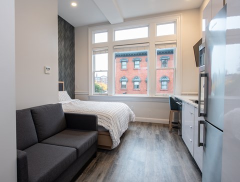 A grey couch sits in a living room with a window overlooking a red brick building.