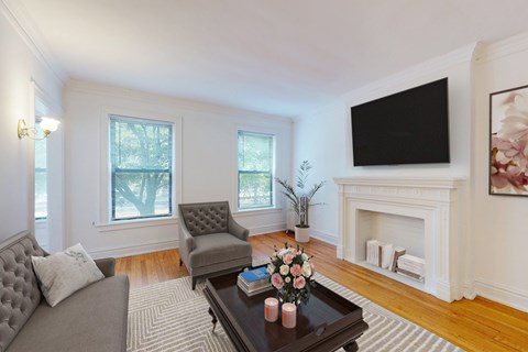 A living room with a grey couch, a grey armchair, a coffee table with flowers and candles, a fireplace, and a flat screen TV mounted on the wall.