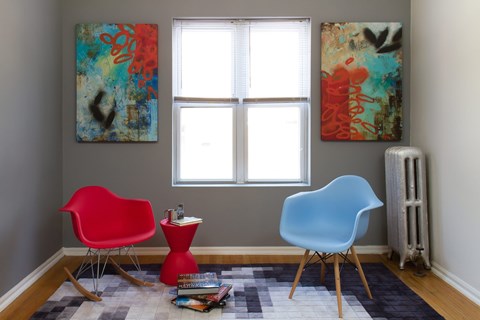 A red chair sits next to a blue chair in a room with two paintings on the wall.