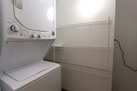 A white washing machine is in a small laundry room with a shelf and a wall outlet.