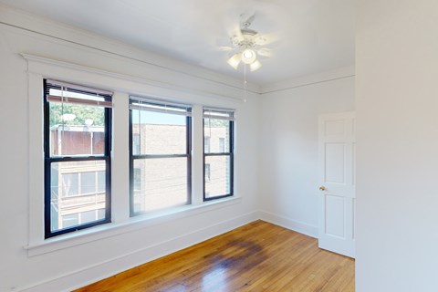 A room with a white door, a window with a view of a building, and a ceiling fan.