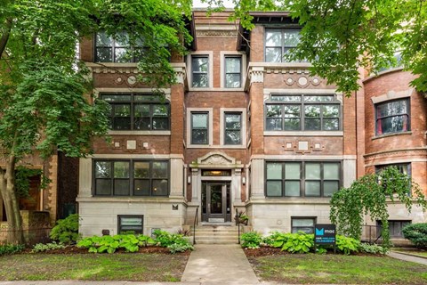 A large, multi-story building with a front entrance and a small garden area in front.