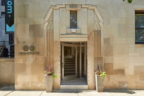 The image shows a building entrance with a stone facade and two potted plants on either side.