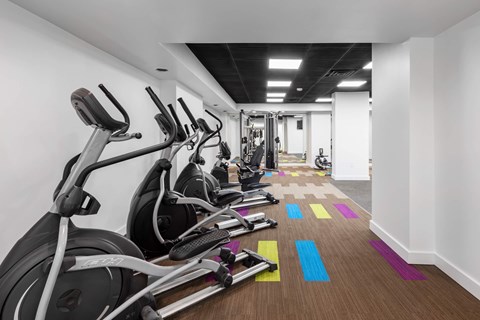 A row of treadmills and exercise bikes are lined up in a brightly lit gym.