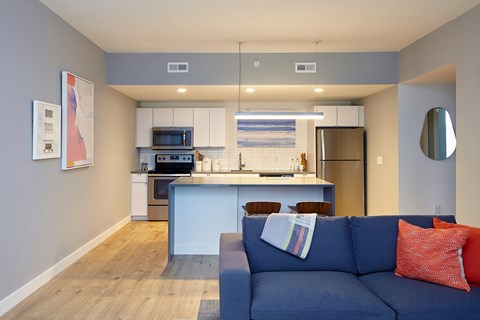 A blue couch with a red pillow is in the foreground of a modern kitchen and living room.
