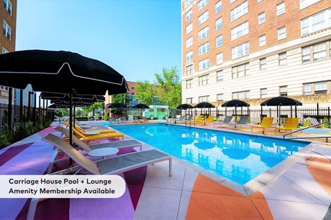 A pool with loungers and umbrellas is surrounded by buildings.