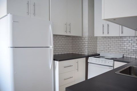 A white refrigerator stands in a kitchen with black countertops and white cabinets.