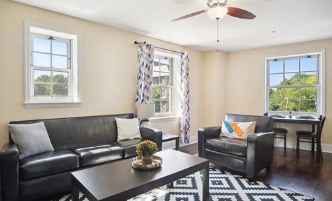A black leather couch sits in a living room with a ceiling fan and a window with a view of trees.
