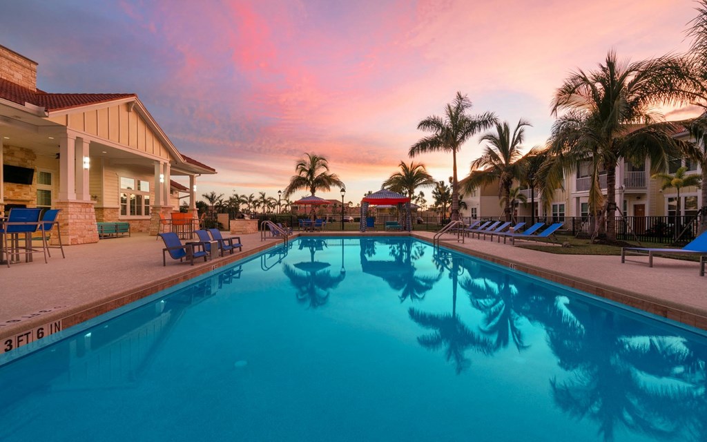a resort style swimming pool with blue chaise lounge chairs and a pink sunset in the background
