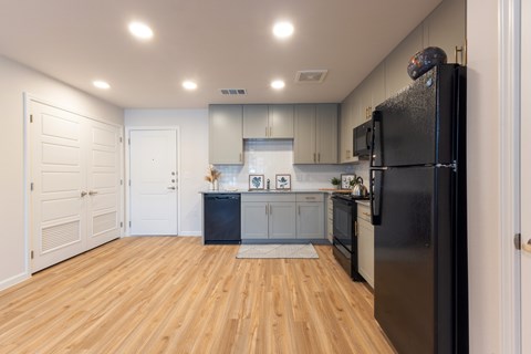 A black refrigerator stands in a kitchen with wooden floors and white walls.