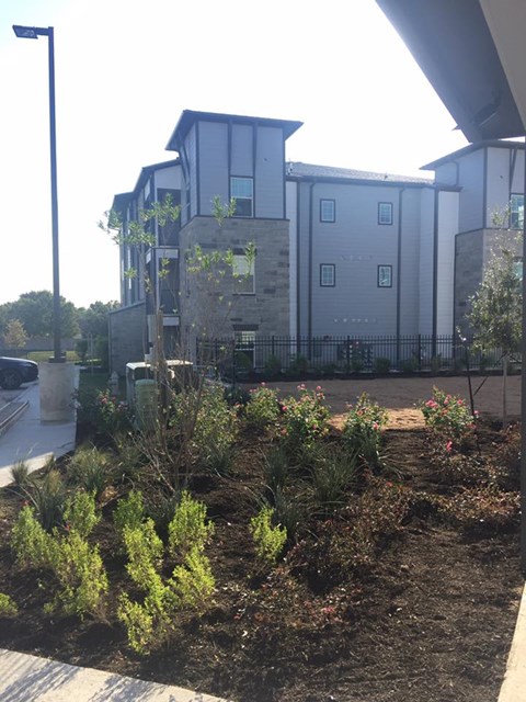 A building with a grey facade is behind a black fence and a garden.